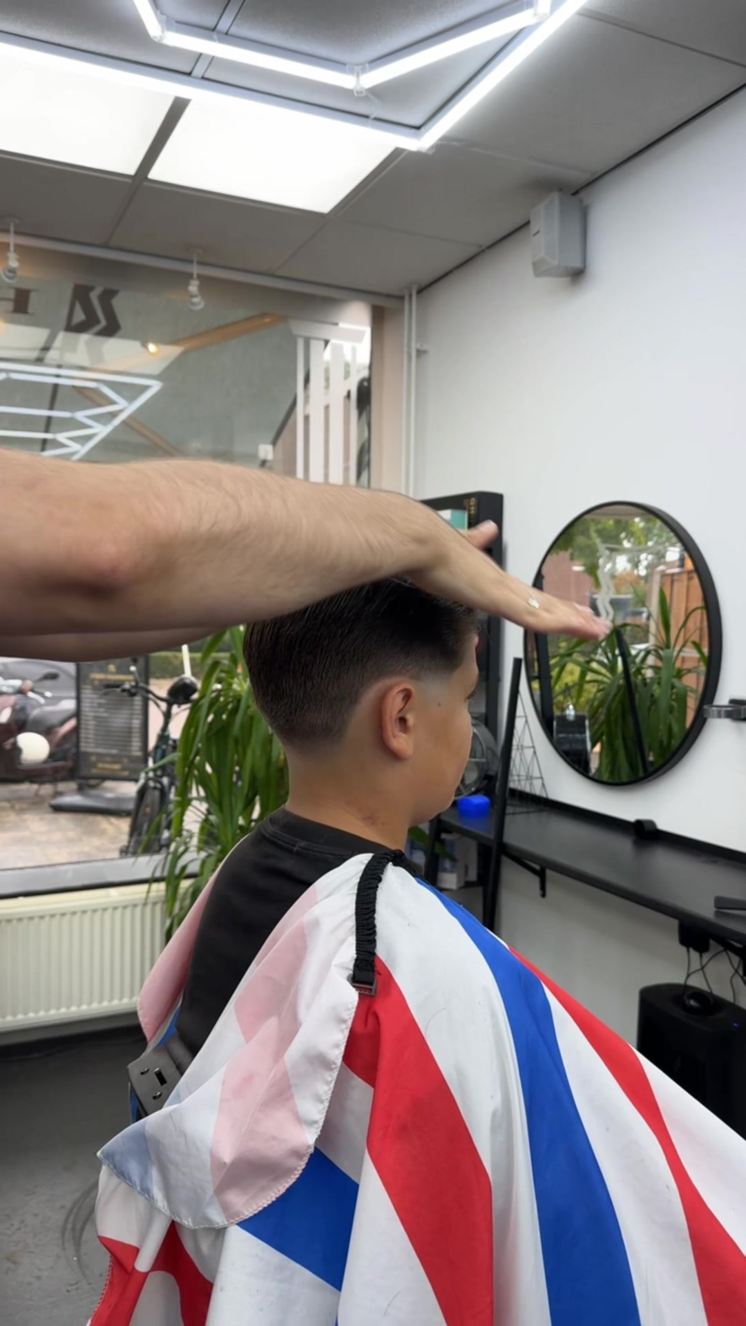 Een jongen wordt geknipt, met de ronde spiegel en de plant van de zaak op de achtergrond